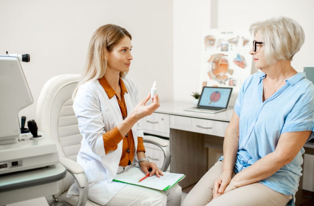 An optician is holding a bottle of pupil-dilating eye drops and explaining their use of it to a  senior woman patient during a comprehensive eye exam
