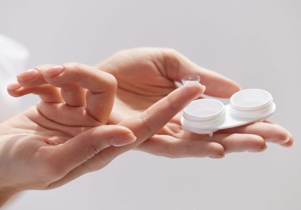 A woman holds a contact lens case in one hand, and she is also holding a contact lens on her index finger.