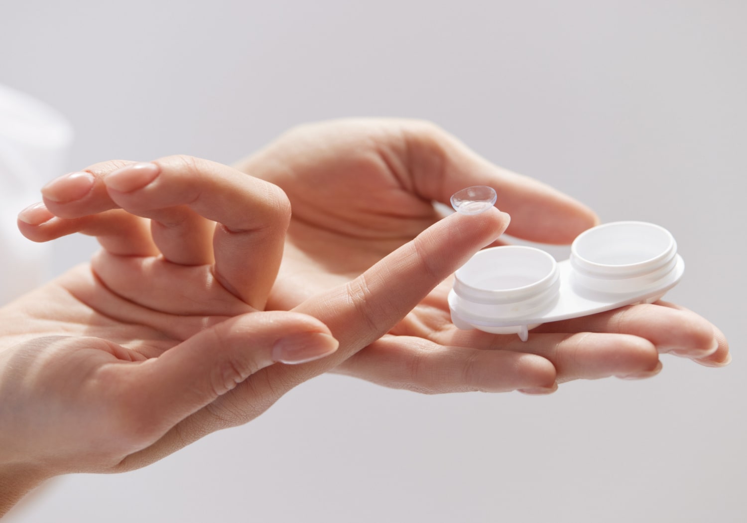 A woman holds a contact lens case in one hand, and she is also holding a contact lens on her index finger.