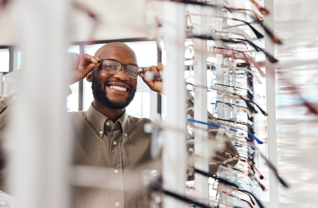 A bearded man trying on a pair of glasses in front of a display rack of frames.