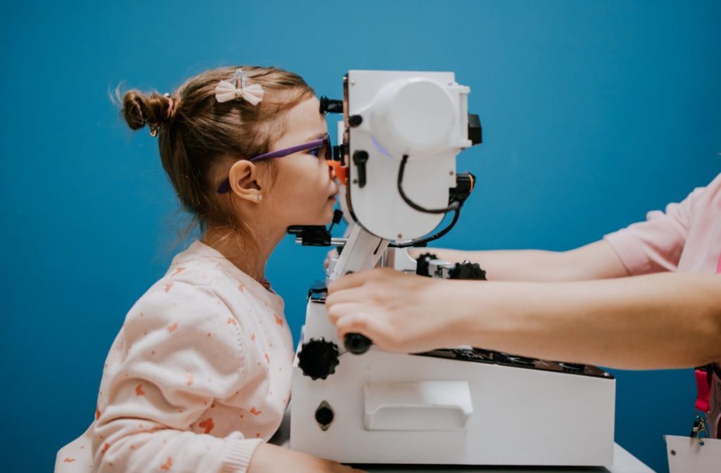 A 5 year old child wearing glasses and a patterned sweater looks through a refractor machine in an optometrist's office.