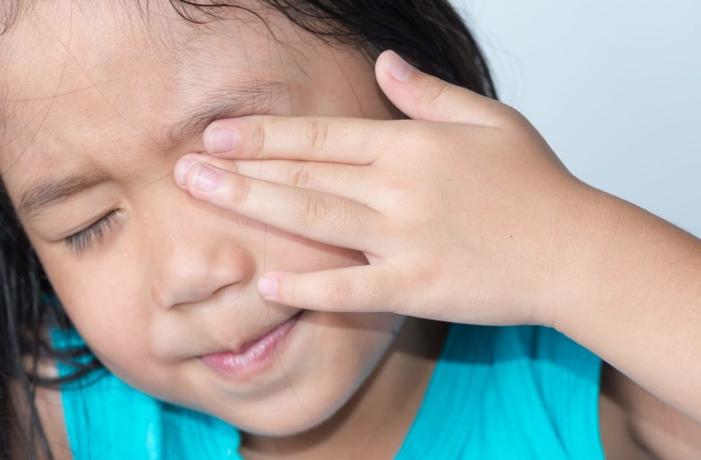 A young child wearing a bright blue shirt rubs their right eye with their hand while squinting their left eye in discomfort.