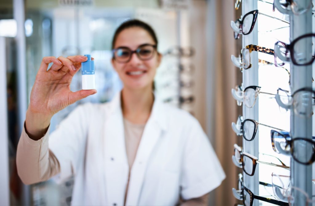 A focused image of an optometrist smiling and holding up a bottle of eye drops in front of a display of eyewear.